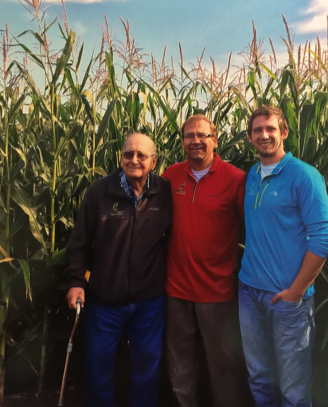 Grandpa with his cane, Dad, and Michael standing together in front of tall tasseled corn